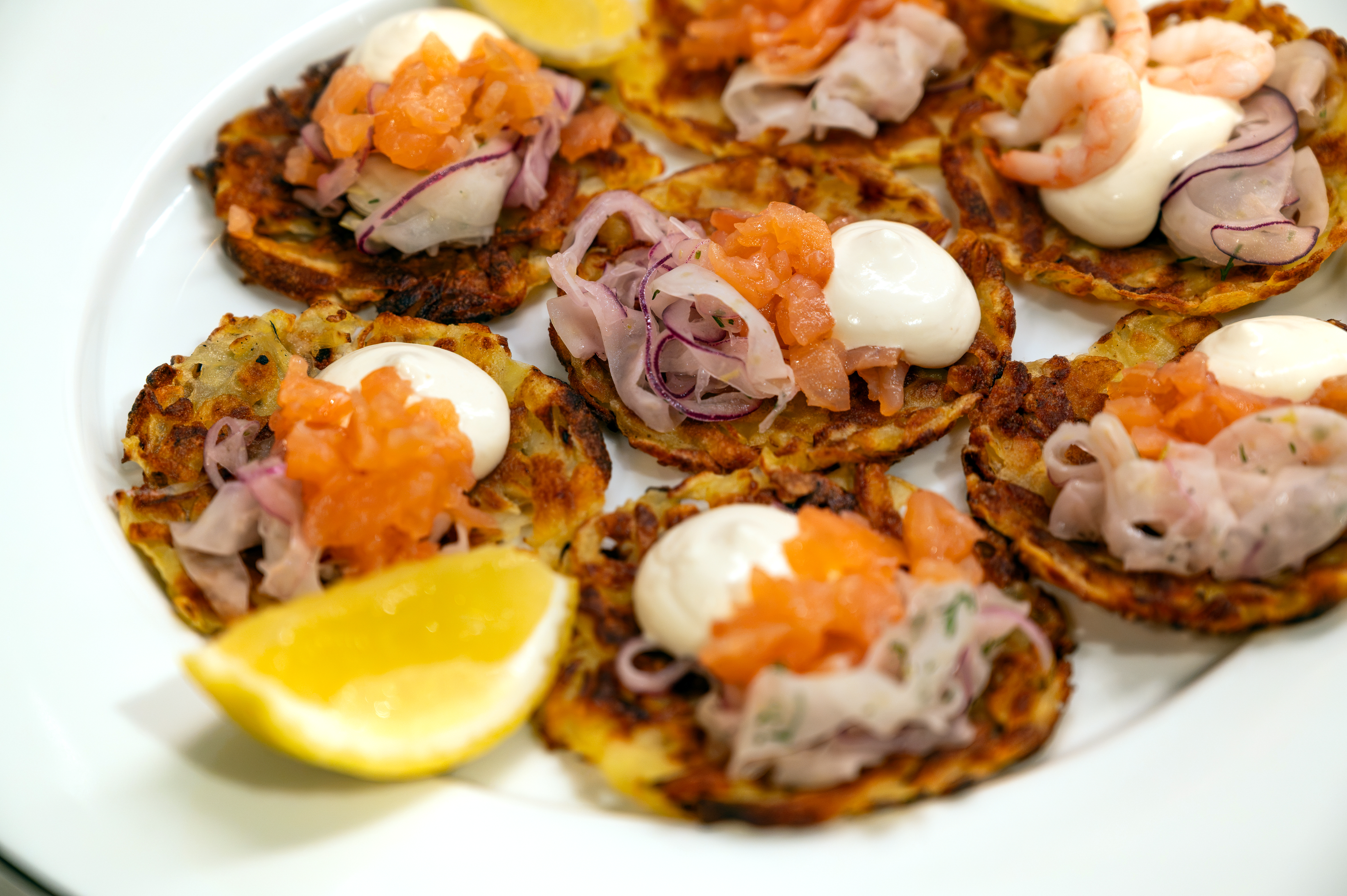 Små krispiga rårakor toppade med lax, räkor, crème fraiche och picklad rödlök på ett vitt fat.